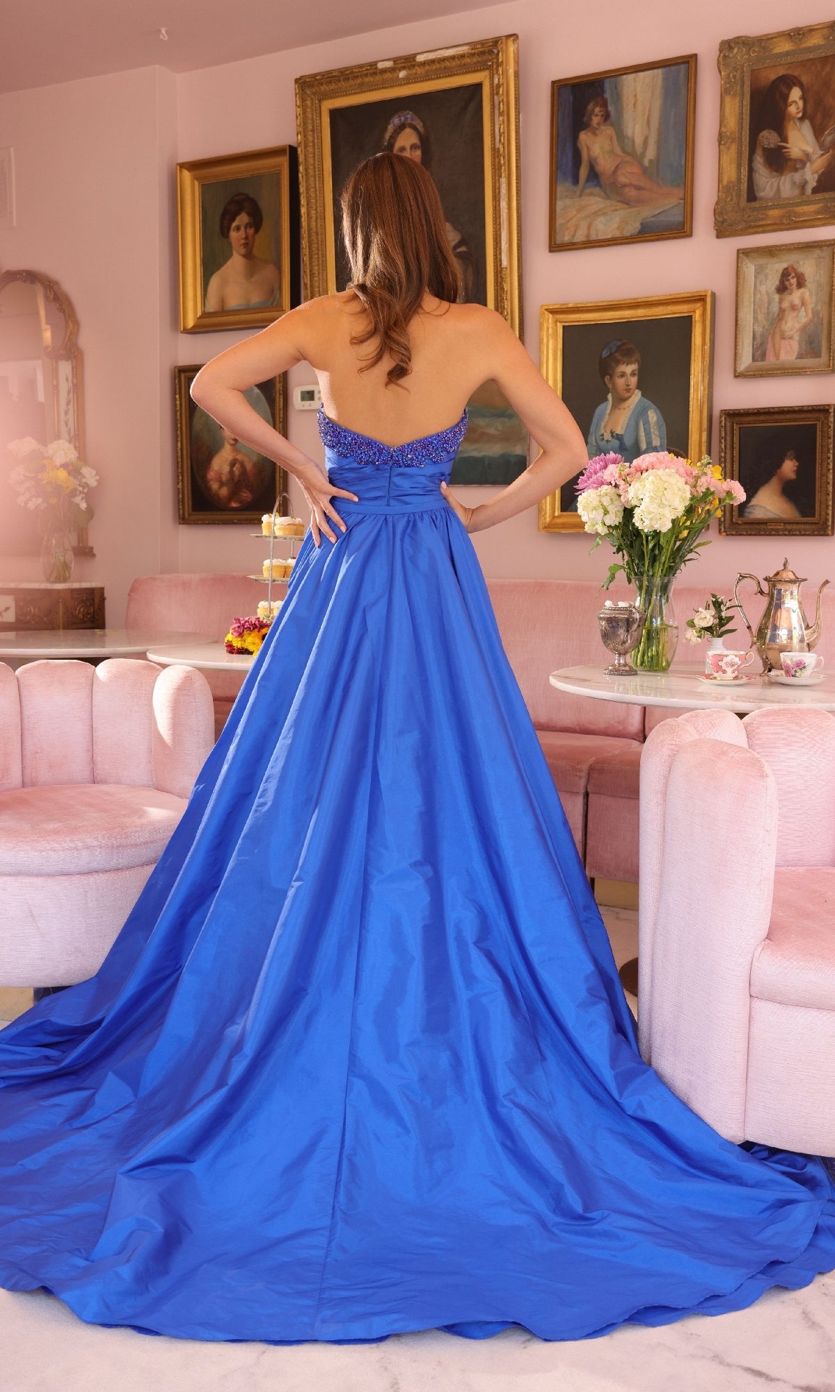 A woman wearing the Ava Presley 40523 long strapless prom dress stands with hands on hips in an elegant room decorated with pink furniture, floral arrangements, and framed portraits.